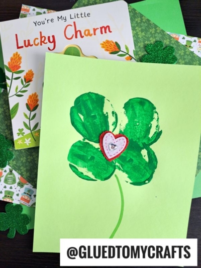 A handmade shamrock card with a heart in the center lies on green paper beside a children’s book titled You’re My Little Lucky Charm. St. Patty’s Day Craft shamrock decorations are also scattered around. @GLUEDTOMYCRAFTS is written at the bottom.