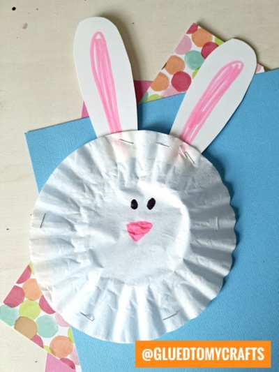 A cute Easter Kid Craft: a bunny face made from a white cupcake liner with pink ears on paper, pink nose, inner ears, and black eyes. Displayed on a light background with colorful paper. Text: @GLUEDTOMYCRAFTS in the corner.