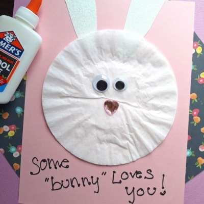 A handmade Easter Kid Craft bunny card made with a coffee filter for the face, googly eyes, a pink heart nose, and white paper ears on pink and floral paper. Text below reads “Some bunny loves you!” and @GLUEDTOMYCRAFTS.