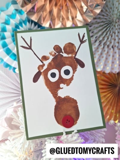A handmade reindeer card with a brown footprint for the face, googly eyes, paper ears, antlers, and a red button nose—perfect for anyone looking for adorable handprint gift ideas. Colorful party fans and the @gluedtomycrafts handle are in the background.