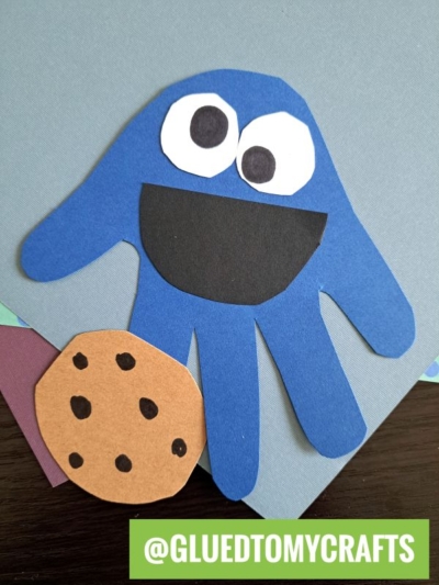 A blue handprint cutout resembles Cookie Monster with large white and black eyes, a big black mouth, and a paper cookie nearby—perfect for fun handprint gift ideas. The handle @GLUEDTOMYCRAFTS is displayed at the bottom in a green box.