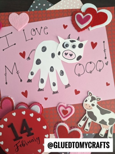 A handmade Valentine’s Day card featuring a cow crafted from a white handprint cutout, decorated with black spots and a pink snout. Surrounded by hearts, it says “I love moo!”—perfect for handprint gift ideas. @GLUEDTOMYCRAFTS is at the bottom.