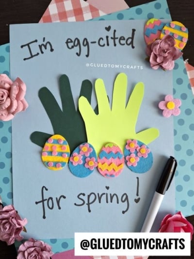 A spring-themed craft featuring two overlapping handprint cutouts (one dark green, one neon yellow-green) resembling grass, with four colorful felt Easter eggs arranged below. The eggs are decorated with patterns like polka dots, flowers, and zig-zags in pink, blue, yellow, and green. The card reads, "I'm egg-cited for spring!" with a small heart above the 'i' in "egg-cited." The craft is displayed on a light blue background with layered patterned papers, rolled paper flowers, and a black marker nearby. The watermark "@GLUEDTOMYGRAFTS" is visible.