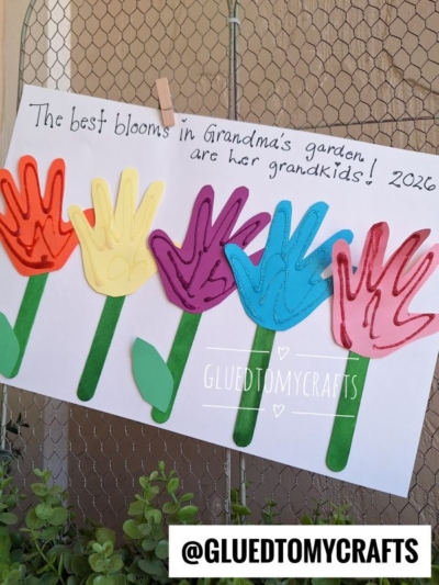 A craft of colorful handprint flowers on sticks is displayed on white paper with the message: The best blooms in Grandma’s garden are her grandkids! 2026. Perfect for those seeking handprint gift ideas. Instagram: @GLUEDTOMYCRAFTS.