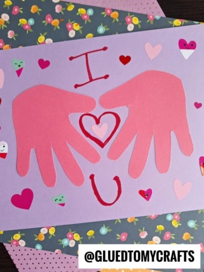 A craft featuring two pink paper hand cutouts forming a heart shape with I ❤️ U in red on a purple background, surrounded by colorful heart stickers—perfect for handprint gift ideas. The handle @GLUEDTOMYCRAFTS appears at the bottom.