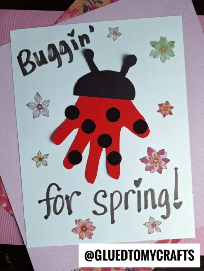 Handmade card with a red handprint ladybug, black paper spots and antennae, surrounded by flower stickers. Perfect as an Easter Kid Craft! Text reads “Buggin’ for spring!” with a heart, and @GLUEDTOMYCRAFTS at the bottom.