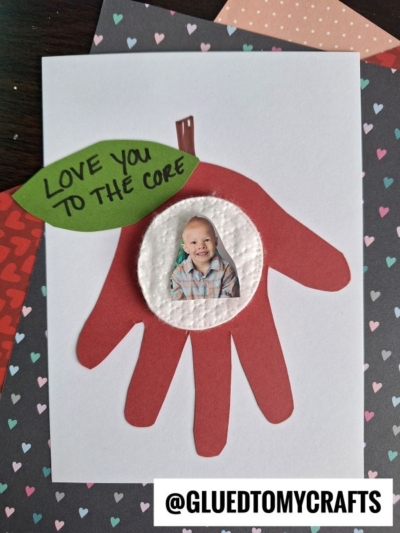 A handmade card, perfect for handprint gift ideas, features a red paper handprint shaped like an apple with a child's photo in the center and a green leaf reading "Love you to the core," set on a heart-patterned background.