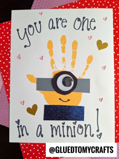A handmade card, perfect for handprint gift ideas, features a yellow handprint decorated as a Minion with a googly eye, gray goggle strip, smiles, and hearts. The card reads, “You are one in a minion!” on a heart-patterned background.