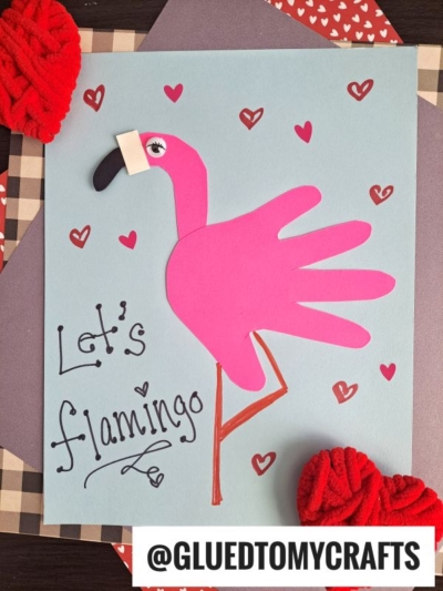 A handmade flamingo crafted from a pink handprint on blue paper, decorated with hearts and the phrase "Lets flamingo." Red yarn and patterned paper frame the artwork—a perfect choice for creative handprint gift ideas. @GLUEDTOMYCRAFTS text at the bottom.