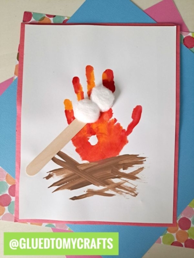 A child’s handprint in red, orange, and yellow forms a campfire, with brown logs, a craft stick, and cotton balls like marshmallows—layered over blue and pink papers. Perfect for creative handprint gift ideas!.