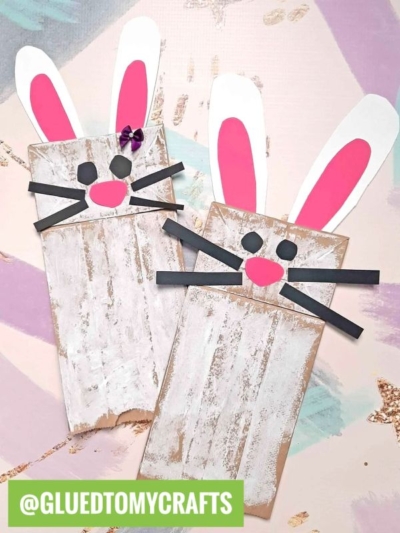 Two Easter Kid Craft paper bag bunnies with pink and white ears, black whiskers, and button noses are displayed on a pastel-colored background. The username @GLUEDTOMYCRAFTS is labeled in green at the bottom.