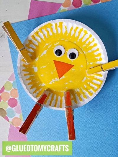 A cute Easter kid craft: a paper plate painted yellow with two googly eyes, an orange triangle beak, and four wooden clothespins as wings and legs. Colorful paper layers create a festive background for this adorable chick.