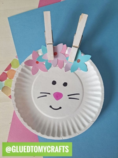 A charming Easter kid craft: a paper plate bunny face with drawn eyes, nose, mouth, and whiskers. Two clothespins topped with paper flowers make cute ears. @GLUEDTOMYCRAFTS is written in green at the bottom.
