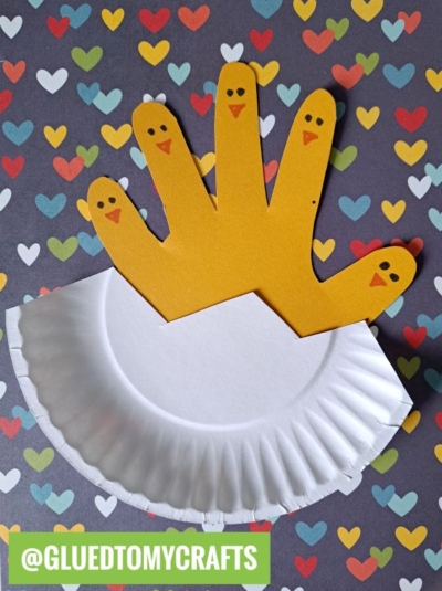 A delightful Easter Kid Craft featuring a yellow handprint cutout with chick faces on each finger, attached to a white half paper plate as an egg, set against a colorful heart-patterned background.