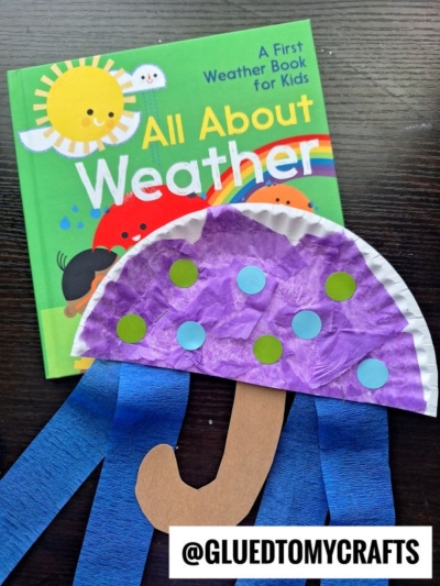 A children's book titled All About Weather sits next to a colorful Easter kid craft—an umbrella made from a paper plate, tissue paper, and construction paper. Both the book and craft rest on a dark surface. @GLUEDTOMYCRAFTS is at the bottom.