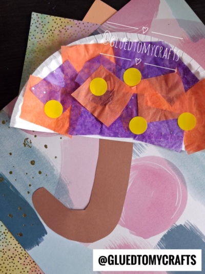 A kids’ craft of a whimsical mushroom, reminiscent of a paper umbrella, made from a paper plate, tissue paper, yellow dots, and brown construction paper, displayed on colorful paper. @GLUEDTOMYCRAFTS is written on the image.