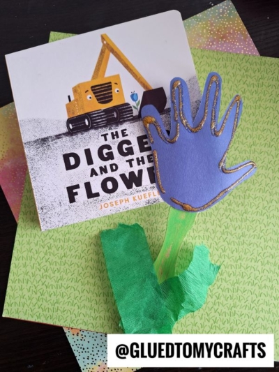 A children’s book titled “The Digger and the Flower” sits behind a hand-shaped blue paper flower craft—perfect for handprint gift ideas—displayed on colorful dotted paper. @GLUEDTOMYCRAFTS is written at the bottom.