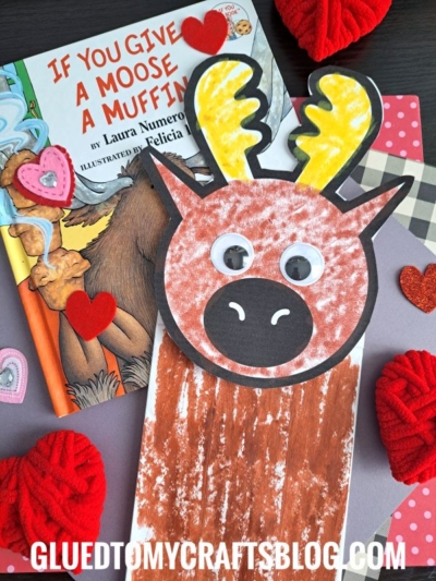 A children’s paper bag moose puppet made from a paper bag and colored paper sits on a table, surrounded by red yarn hearts, a book titled If You Give a Moose a Muffin, and other heart-shaped decorations.