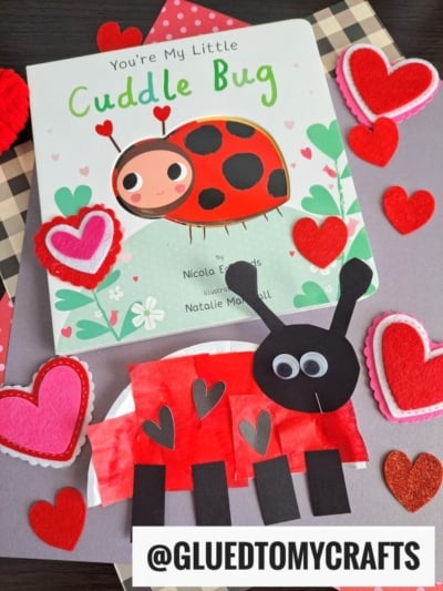 A children’s book titled “You’re My Little Cuddle Bug” is surrounded by heart-shaped decorations and a handmade paper plate ladybug craft, made with paper, felt, and construction paper. Instagram handle @GLUEDTOMYCRAFTS is at the bottom.