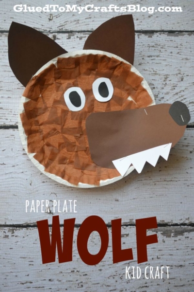 A paper plate wolf craft made with brown paper, white teeth, and a black nose, displayed on a wooden surface. This adorable paper plate wolf project is easy for kids to make! Text reads Paper Plate Wolf Kid Craft and GluedToMyCraftsBlog.com.