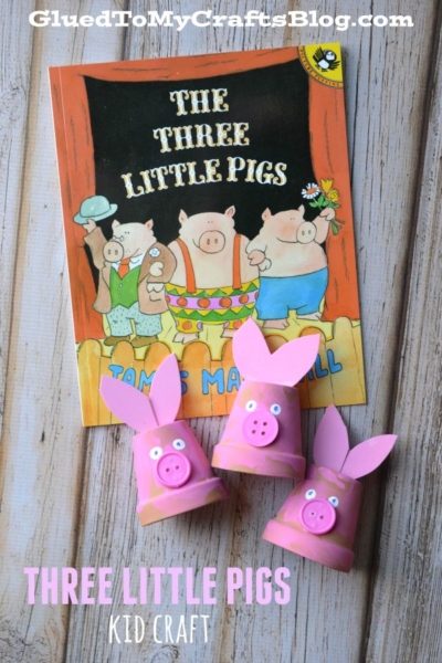 Children’s book “The Three Little Pigs” with three pink pig crafts made from upside-down terra cotta pots, pink ears, and button noses, displayed on a wooden surface.