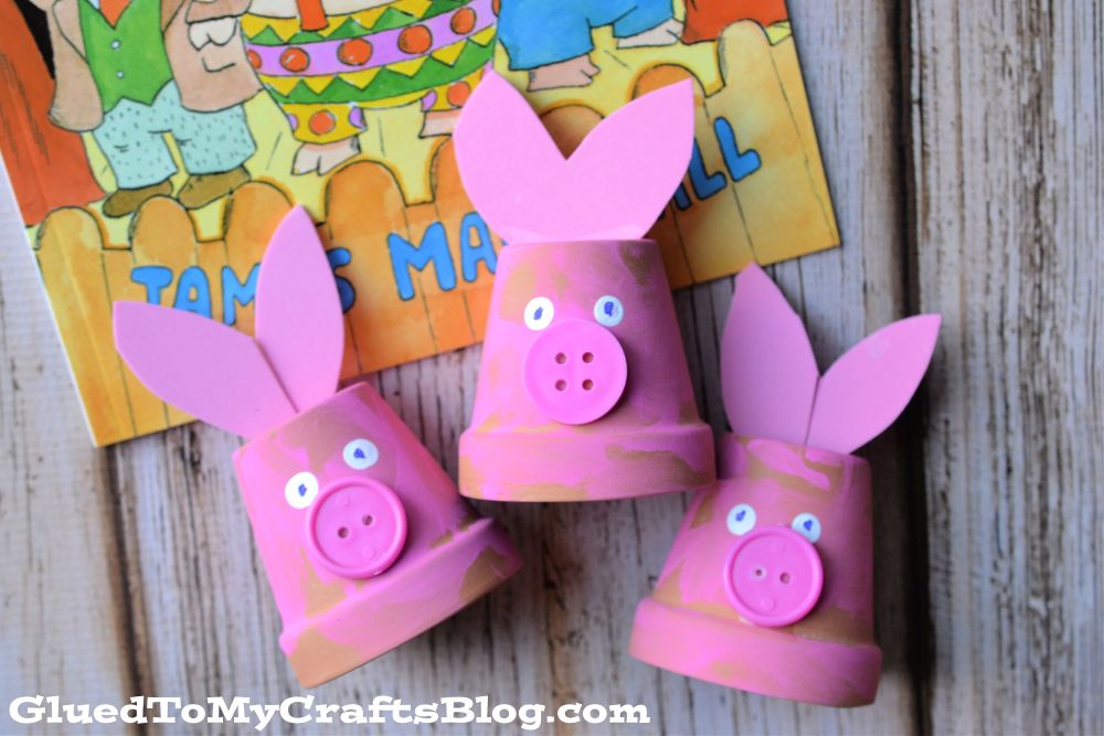 Three adorable pig crafts, inspired by the classic tale of the three little pigs, are made from small pink terra cotta pots, pink paper ears, googly eyes, and button noses. Displayed in front of a colorful children’s book, the image includes the watermark GluedToMyCraftsBlog.com.