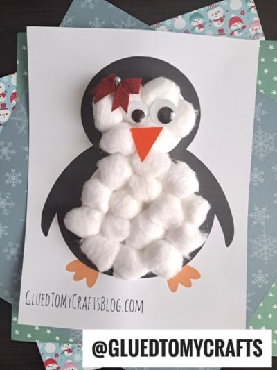 A handmade cotton ball penguin craft features a black and white paper penguin decorated with cotton balls on its body, orange paper beak and feet, googly eyes, and a red bow on its head, displayed on winter-themed paper.