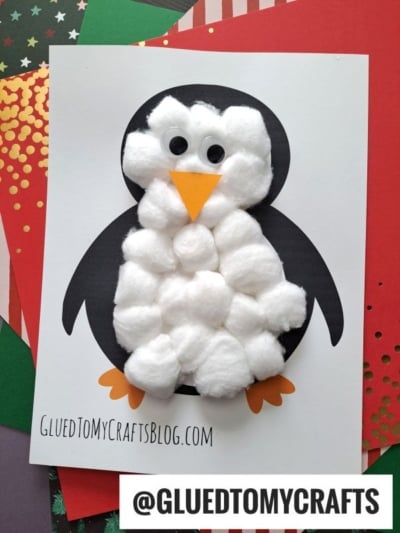 A handmade cotton ball penguin craft featuring a fluffy body made from cotton balls, an orange paper beak and feet, googly eyes, all showcased on a white card with @GLUETOMYCRAFTS and colorful paper in the background.