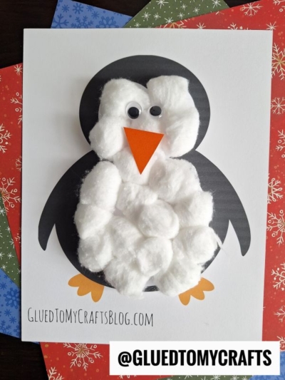 A cotton ball penguin craft made from black and white paper, orange feet and beak, googly eyes, and cotton balls for the body, displayed on a festive snowflake-patterned background.