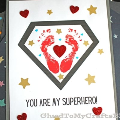 A handmade footprint superhero card features red baby footprints inside a gray superhero emblem, surrounded by gold stars and red glitter hearts. The text at the bottom reads, “YOU ARE MY SUPERHERO!”.