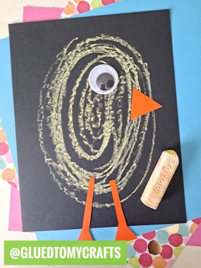 A charming Easter Kid Craft featuring a chick with a chalk-drawn oval body on black paper, a large googly eye, orange paper beak and legs, plus a piece of chalk beside it. @GLUEDTOMYCRAFTS is written at the bottom.