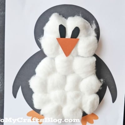 A cotton ball penguin craft made with black paper, a belly covered in cotton balls, orange paper beak and feet, and black paper eyes, displayed on a white background.