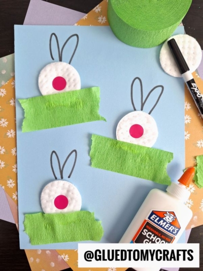 A blue sheet displays three Easter kid craft bunny creations made from cotton pads with pink centers, drawn ears, and green crepe paper grass. Elmer’s glue, a black marker, and pastel papers surround the scene. Text reads “@GLUETOMYCRAFTS.”.
