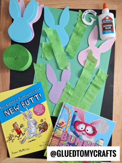 A flat lay Easter Kid Craft featuring bunny-shaped cutouts, green tissue strips, a pompom, glue, and two Easter bunny-themed books is arranged on a black surface. The handle @GLUEDTOMYCRAFTS appears at the bottom.