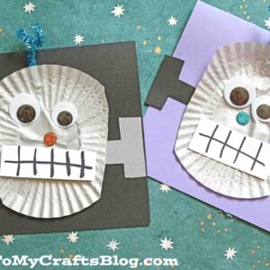 Two Cupcake Liner Robots decorated as Frankenstein monster faces are made with cupcake liners, googly eyes, and paper, then glued to black and purple squares. Each robot features pipe cleaner hair, a drawn mouth, and square bolts on the sides.