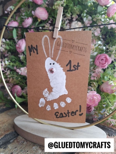 A handmade Easter card, perfect as an Easter Kid Craft, features a white painted bunny made from a child's footprint and hangs on a string. "My 1st Easter!" is written on the card, with pink flowers and greenery blurred in the background.