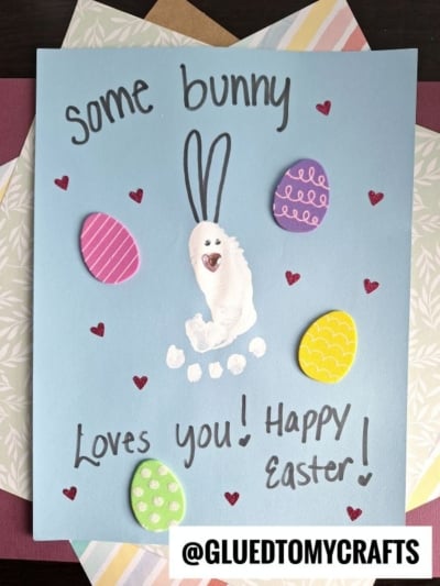 A blue Easter card features a white bunny made from a child’s footprint, surrounded by foam Easter eggs and red hearts. This adorable Easter Kid Craft reads, "Some bunny loves you! Happy Easter!" with @GLUEDTOMYCRAFTS at the bottom.
