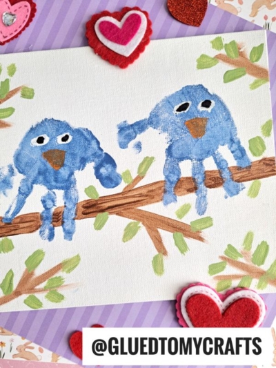 A painting of two blue birds made from handprints sits on a tree branch, surrounded by painted leaves. This Easter Kid Craft is decorated with red and pink felt hearts and has the text @GLUETOMYCRAFTS at the bottom.