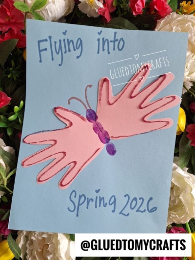A handmade Easter Kid Craft butterfly made with two pink handprints on blue paper, with Flying into Spring 2026 written in blue and surrounded by colorful flowers. @GLUEDTOMYCRAFTS appears at the bottom.