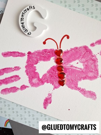 A pink butterfly made from handprints on white paper, with red heart-shaped gems as its body. This charming Easter Kid Craft features a “GluedToMyCrafts” label above and “@GLUEDTOMYCRAFTS” at the bottom.