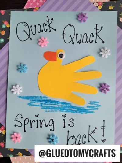 A handmade Easter kid craft card features a yellow paper duck made from a traced handprint, googly eye, orange beak, blue crayon water, decorative flowers, and the text Quack Quack Spring is back! with @GLUEDTOMYCRAFTS.