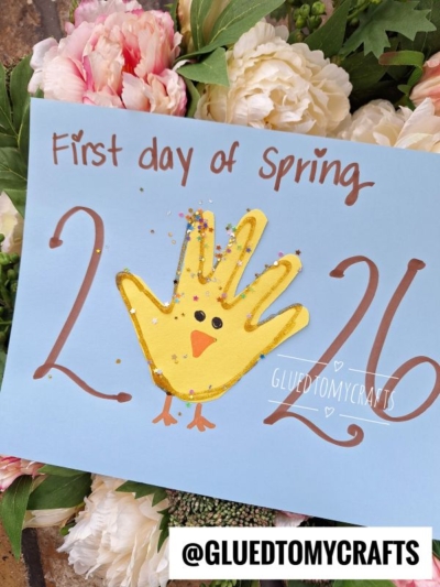 A blue paper reads First day of Spring 2026 with a yellow handprint chick drawing between the numbers. This Easter kid craft is decorated with glitter, surrounded by flowers, and features @GLUEDTOMYCRAFTS at the bottom.