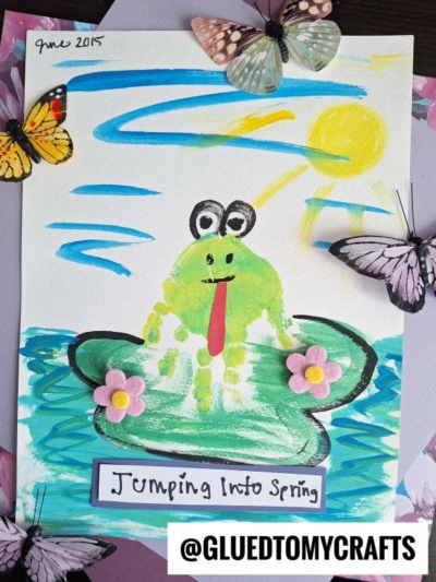 A colorful children’s painting shows a green frog with googly eyes and a red tongue sitting on a lily pad with two pink flowers, under a bright sun, with decorative butterflies attached. This cheerful Easter Kid Craft reads “Jumping Into Spring.”.