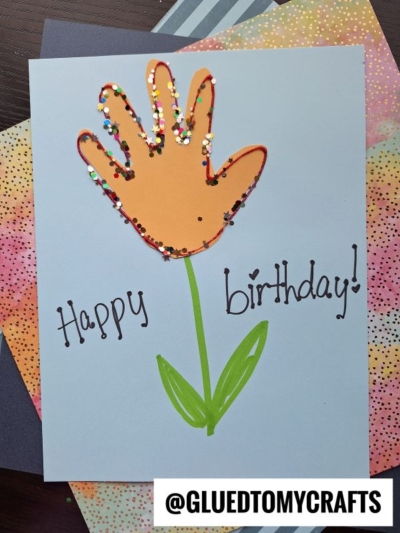 A handmade birthday card featuring an orange handprint—perfect for handprint gift ideas—outlined in red and decorated with colorful glitter to form a flower on a green stem, with "Happy birthday!" written in black marker.