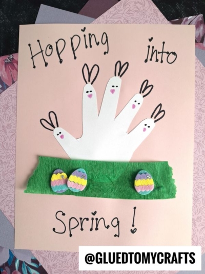 A fun Easter Kid Craft featuring a white handprint with drawn bunny faces and ears, green crepe paper grass, and colorful Easter egg cutouts on a pink background. Text reads Hopping into Spring! and @GLUEDTOMYCRAFTS.