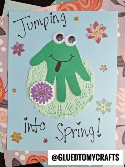 A fun Easter Kid Craft featuring a green handprint frog with googly eyes and a smile, sitting on a paper doily. The playful text says Jumping into Spring! surrounded by cheerful floral decorations.