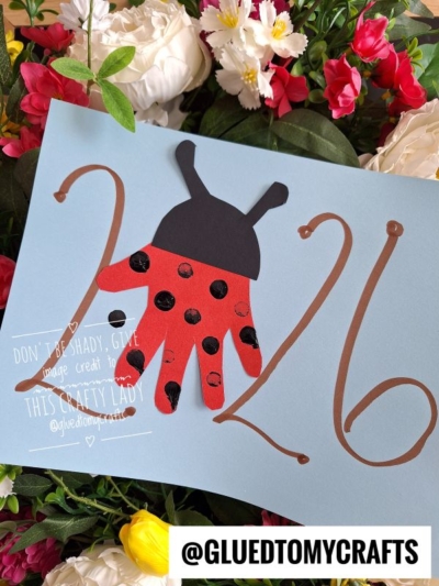 A blue Easter Kid Craft card with 2026 in brown, featuring a red and black ladybug handprint as the zero. Surrounded by vibrant pink, white, and yellow flowers. @GLUEDTOMYCRAFTS is written at the bottom.