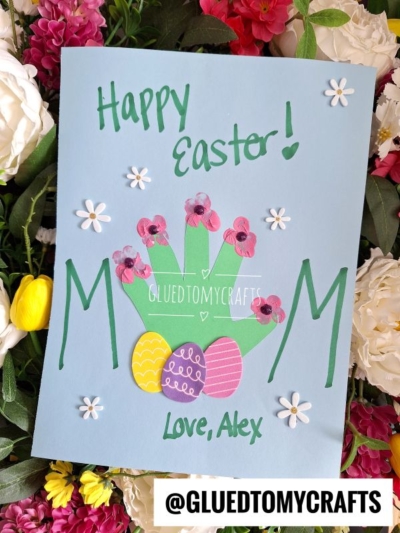 A blue Easter Kid Craft card with “Happy Easter!” on top, featuring a green handprint decorated with pink flowers, “MOM” in green letters, colorful Easter eggs, and “Love, Alex” at the bottom. Flowers bloom in the background.