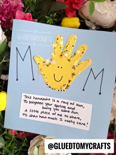 A handmade Mothers Day card featuring a yellow handprint, star confetti, and a smiley face, surrounded by colorful flowers. Perfect as an Easter Kid Craft, black letters spell MOM and a note reads, “This handprint is a ray of sun...”.