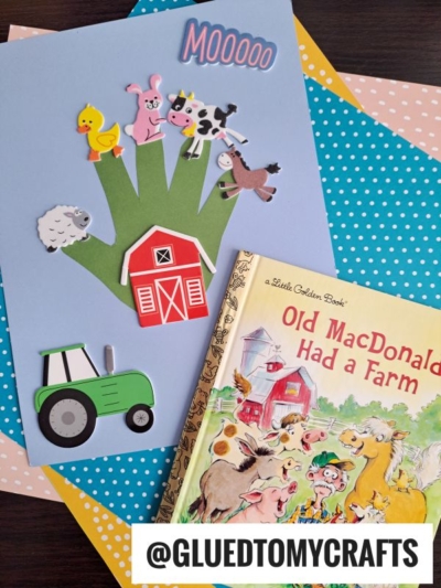 A paper craft of a green hand decorated with farm animal stickers, a red barn, and a green tractor sits next to the book Old MacDonald Had a Farm on colorful polka dot papers—perfect for creative handprint gift ideas. @GLUEDTOMYCRAFTS is labeled at the bottom.
